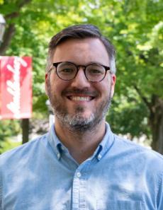 A man smiles, standing in front of a Muhlenberg College sign and trees.